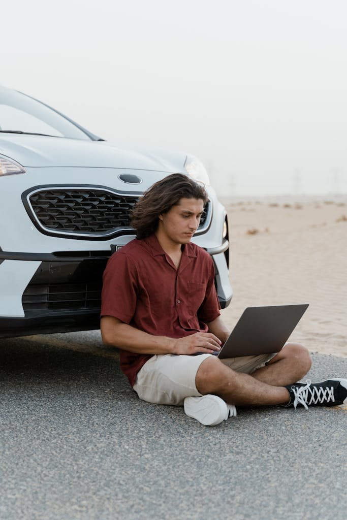 Man sitting by car on desert road with laptop, embracing remote work lifestyle.